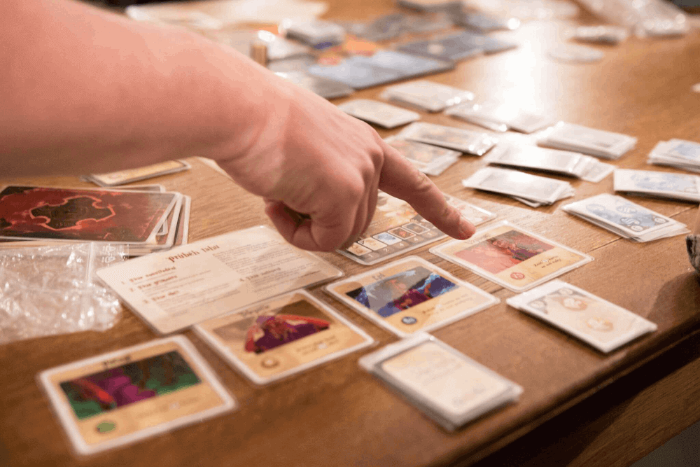 stůl plný herních karet a ukazováček ruky hráče mířící na jednu z aktivních herních kartiček vyloženou na stole